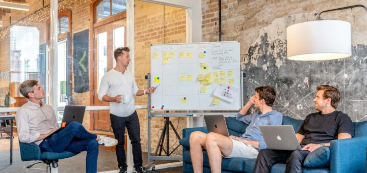 three men sitting while using laptops and watching man beside whiteboard
